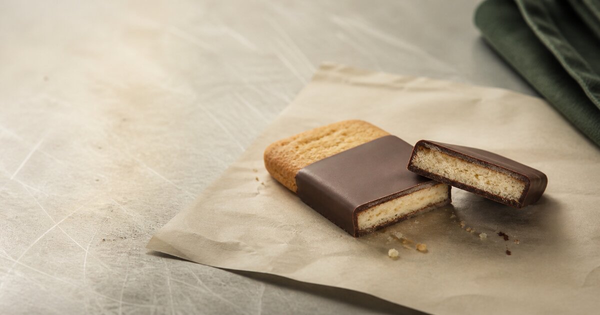 A cut chocolate-coated biscuit showing a thin coating around pale crumb on parchment at a bakery bench